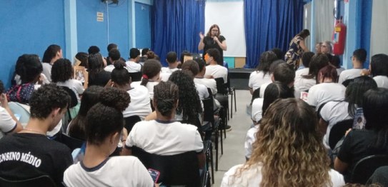 Descrição da imagem: Cinco alunos(as), três meninas e dois meninos, estão em pé e fazem fila par...