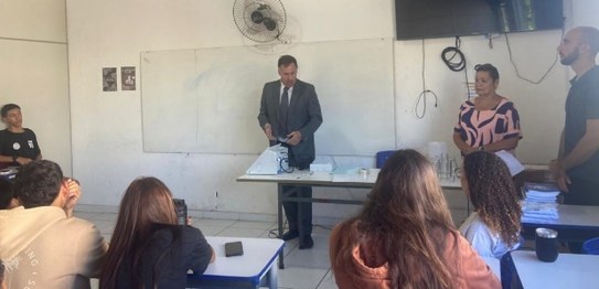 No centro da sala, o juiz veste um terno preto e conversa com os alunos, que estão sentados em c...