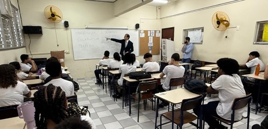 Auditório com cortina vermelha ao fundo, onde um homem está à frente, conduzindo uma apresentaçã...