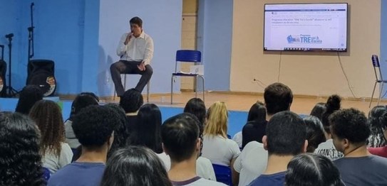 Descrição da imagem: O juiz veste um terno preto e segura um microfone em cima do palco. As(os) ...