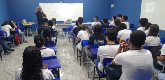 Um palestrante fala para jovens sentados em cadeiras azuis em um auditório simples, com bandeira...