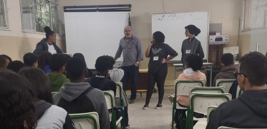 No centro da sala, o juiz veste um terno preto e conversa com os alunos, que estão sentados em c...