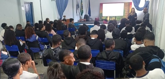 Descrição da imagem: O juiz veste um terno preto e segura um microfone em cima do palco. As(os) ...