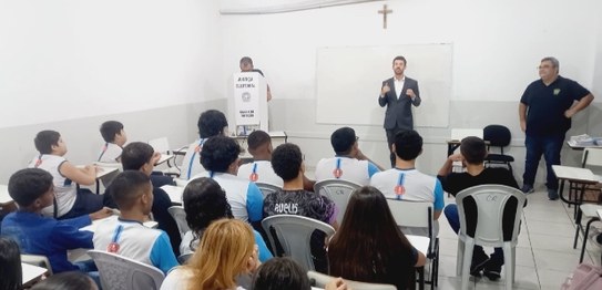 Descrição da imagem: Em pé, o juiz veste um terno e está de frente para os(as) alunos(as). Os(as...