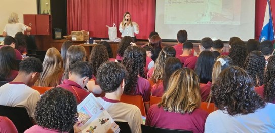 No centro da sala, o juiz veste um terno preto e conversa com os alunos, que estão sentados em c...