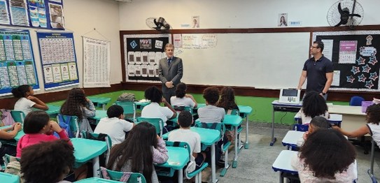 Um palestrante fala para jovens sentados em cadeiras azuis em um auditório simples, com bandeira...