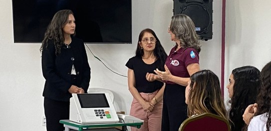 Numa sala, sentados, estudantes assistem à palestra da juíza eleitoral. Ela está de pé na frente...