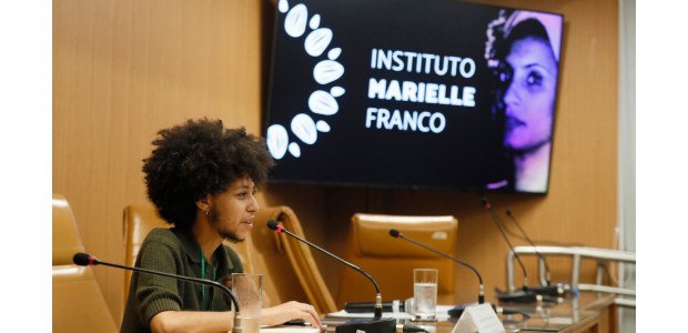 Um homem está sentado de perfil, falando ao microfone em uma mesa de conferência. Ele veste uma ...