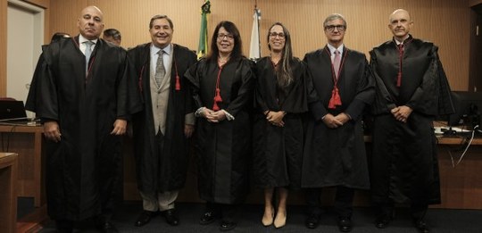 A imagem mostra seis pessoas em pé, todas vestindo togas pretas com detalhes em vermelho, o que ...