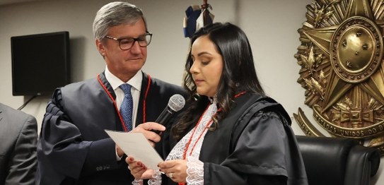 Manoela Dourado, mulher branca, cabelos longos e pretos, vestindo toga, fala ao microfone; o pre...