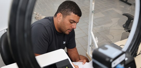 foto do atendimento biométrico no Cipop-Rua. À esquerda, um homem, sentado atrás de um acrílico ...