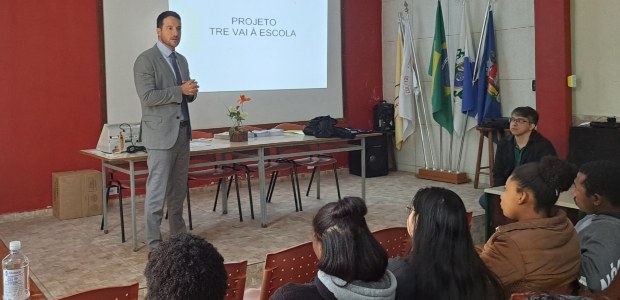 Magistrado,  usando terno cinza, conduz uma palestra para jovens em uma sala com telão, onde se ...