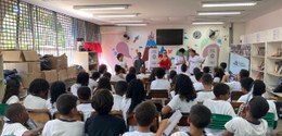 Descrição da imagem: numa sala de aula, a palestrante, ladeada por  quatro estudantes, fala para...