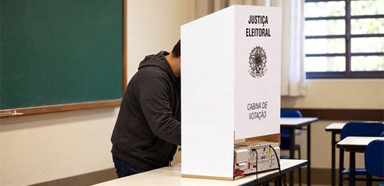 Em primeiro plano, sobre uma mesa escolar, encontra-se uma cabina de votação eleitoral em papelã...