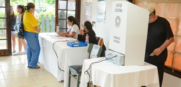 A imagem mostra um local de votação da Justiça Eleitoral. Em primeiro plano, há uma cabine de vo...
