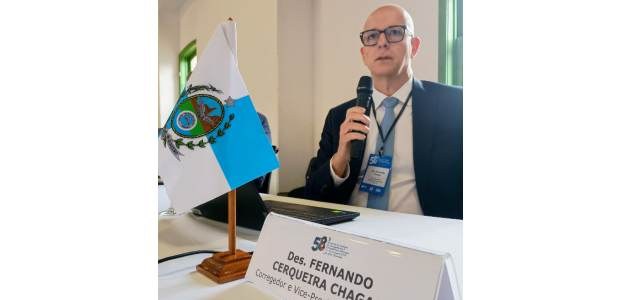 Desembargador Fernando Chagas está sentado à mesa, falando ao microfone. Ele usa óculos e está v...