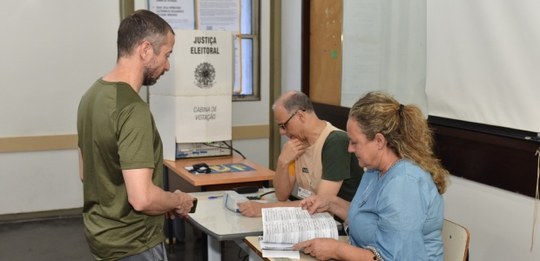 Em primeiro plano, um homem está diante da mesa de atendimento, enquanto duas pessoas que atuam ...