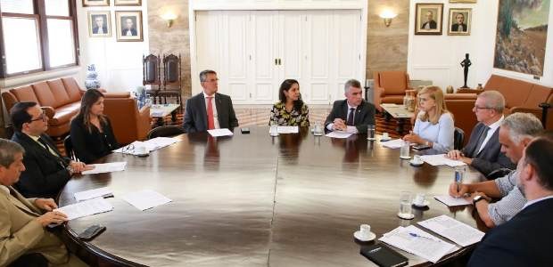 Grupo de 10 autoridades reunidas em torno de uma grande mesa redonda em sala institucional , ten...