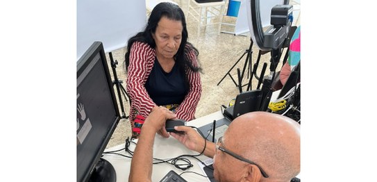 A imagem mostra uma mulher sendo atendida por um servidor da Justiça Eleitoral, que realiza a co...