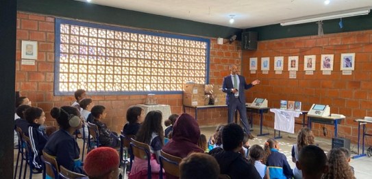 Alunos sentados assistem palestra feita por homem de terno, que aponta para duas urnas eletrônic...