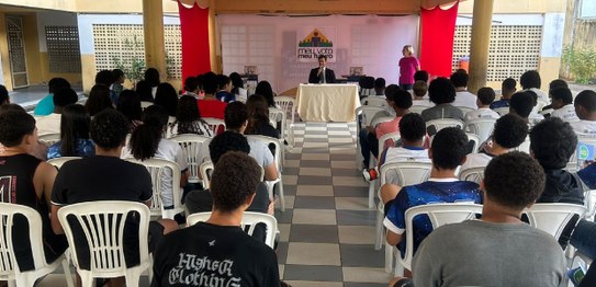 A imagem mostra um ambiente de sala de aula com um quadro branco ao fundo e estudantes sentados....