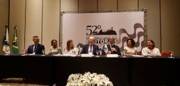 Em primeiro plano, sentados, sete participantes da mesa de abertura do evento. Ao centro, o pres...