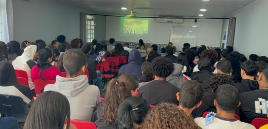 Em uma sala ampla e iluminada, quatro palestrantes de pé ao lado de bandeiras apresentam uma ati...