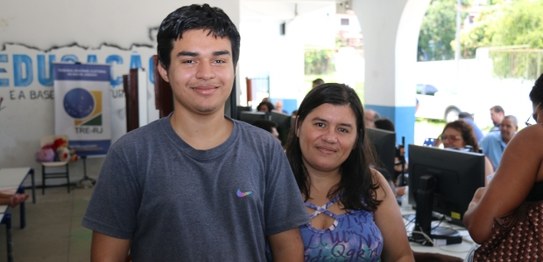 Jovem moreno, com cabelo curto e escuro, sorri ao lado da mãe, com cabelo longo. Eles posam tend...