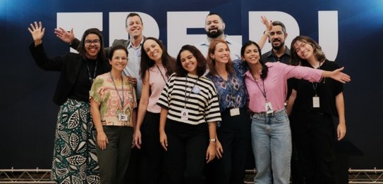 A imagem mostra um grupo de dez pessoas posando para uma foto em frente a um painel instituciona...