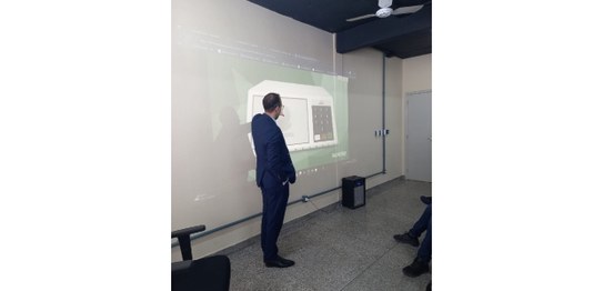 Alunos sentados assistem palestra feita por homem de terno, que aponta para duas urnas eletrônic...