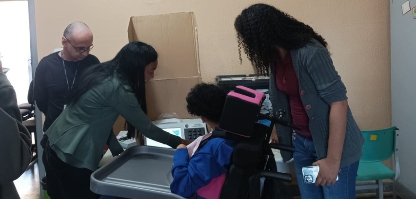 Ao centro da imagem, uma aluna cadeirante vota na urna eletrônica com a ajuda de uma professora....