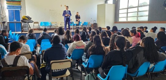 Descrição da imagem: O juiz veste um terno preto e segura um microfone em cima do palco. As(os) ...