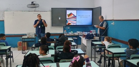 Alunos sentados assistem palestra feita por homem de terno, que aponta para duas urnas eletrônic...