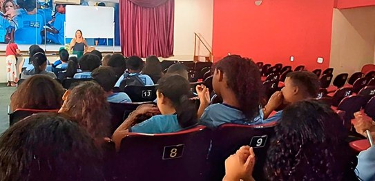 No centro da sala, o juiz veste um terno preto e conversa com os alunos, que estão sentados em c...