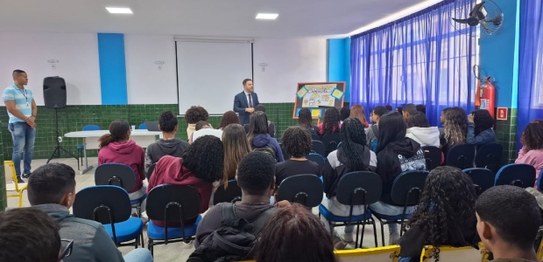 Auditório com cortina vermelha ao fundo, onde um homem está à frente, conduzindo uma apresentaçã...