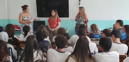 No centro da sala, o juiz veste um terno preto e conversa com os alunos, que estão sentados em c...