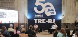 foto ampla do evento, com um palestrante e mediador sentados em cadeiras, tendo ao fundo painel ...