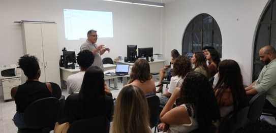 A imagem mostra um homem palestrando para um grupo de jovens sentados em uma sala com computador...