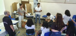 sala de aula com dois palestrantes à frente gesticulando, ao lado de urna eletrônica