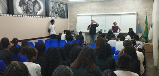 Numa sala de aula, o palestrante gesticula, falando no microfone, tendo ao fundo uma lousa branc...