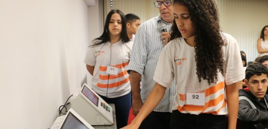 Estudante, de pé, participa de eleição simulada. Ela está apertando uma das teclas da urna eletr...