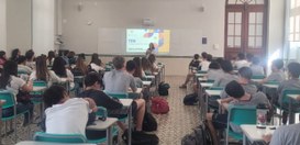 Uma sala de aula cheia de estudantes, sentados em carteiras verdes, assistindo a uma palestra mi...
