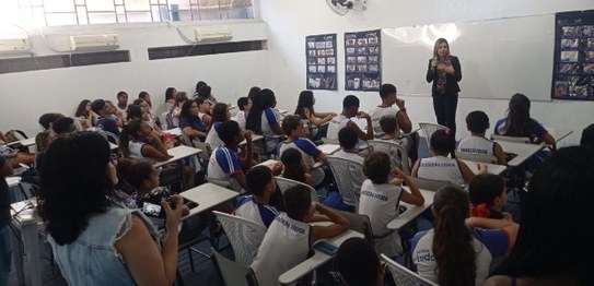 No centro da sala, o juiz veste um terno preto e conversa com os alunos, que estão sentados em c...