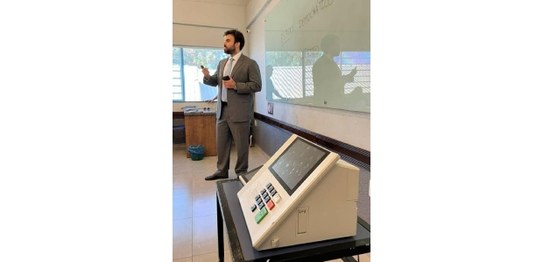 Alunos sentados assistem palestra feita por homem de terno, que aponta para duas urnas eletrônic...