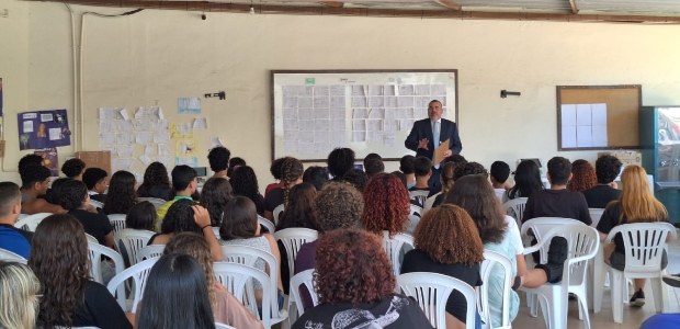 Homem em pé, falando para um grupo de estudantes sentados em cadeiras brancas de plástico, em um...
