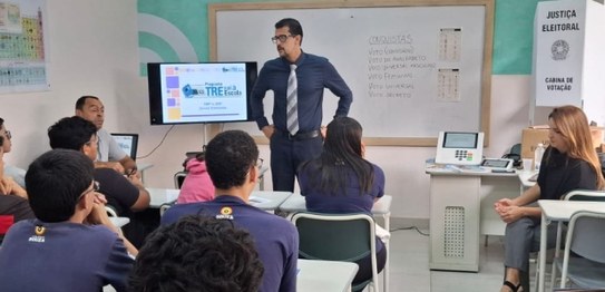 Auditório com cortina vermelha ao fundo, onde um homem está à frente, conduzindo uma apresentaçã...