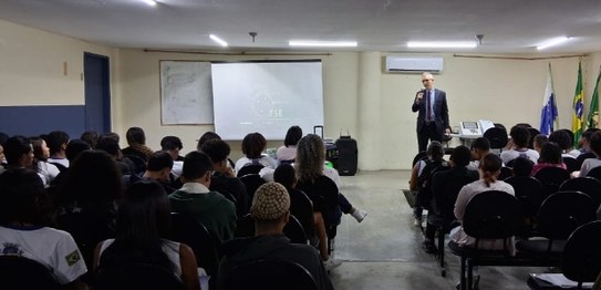 Um palestrante fala para jovens sentados em cadeiras azuis em um auditório simples, com bandeira...