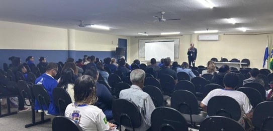 Auditório com cortina vermelha ao fundo, onde um homem está à frente, conduzindo uma apresentaçã...