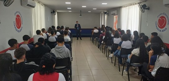 Um palestrante fala para jovens sentados em cadeiras azuis em um auditório simples, com bandeira...