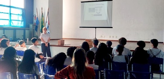 Auditório com cortina vermelha ao fundo, onde um homem está à frente, conduzindo uma apresentaçã...